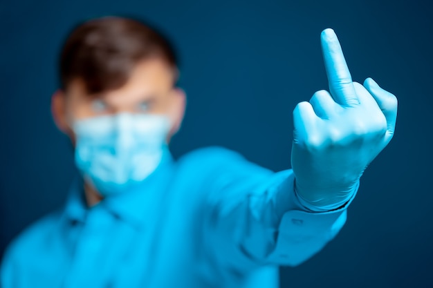 Ein Arzt in einer medizinischen Maske und Handschuhen in blauer Uniform zeigt den Mittelfinger.