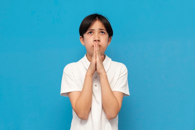 Ein asiatischer Junge betet mit gefalteten Händen vor sich und bittet in einem weißen T-Shirt auf blauem Hintergrund