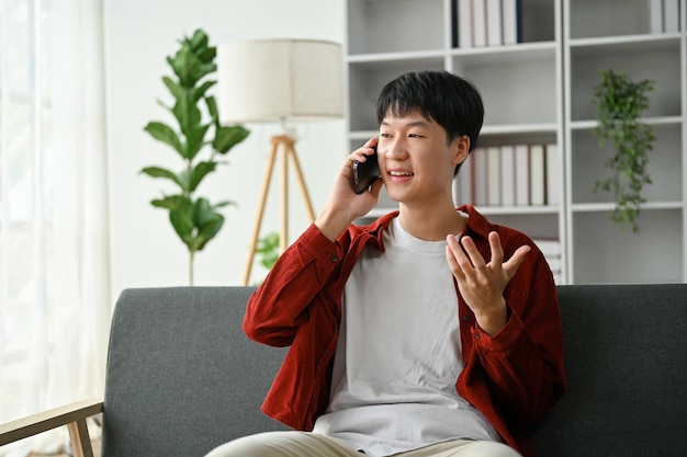 Ein asiatischer Mann, der mit seinem Freund telefoniert, während er auf seinem Sofa sitzt