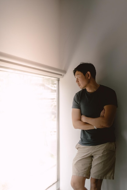 Foto ein asiatischer mann, der sich an eine weiße wand lehnt, in einem marineblauen t-shirt und khaki-shorts und aus den fenstern schaut