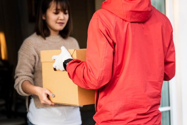 Ein asiatischer Mann in roter Uniform, der einem weiblichen Kunden vor dem Haus ein Paket übergibt. Ein Postbote und ein Expresszustelldienst liefern Pakete während der Covid19-Pandemie.