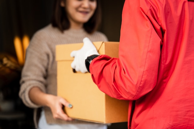 Ein asiatischer Mann in roter Uniform, der einem weiblichen Kunden vor dem Haus ein Paket übergibt. Ein Postbote und ein Expresszustelldienst liefern Pakete während der Covid19-Pandemie.