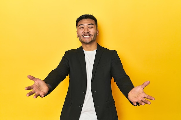 Ein asiatischer Mann in schwarzer Geschäftsjacke auf einem gelben Studio zeigt einen willkommenen Ausdruck