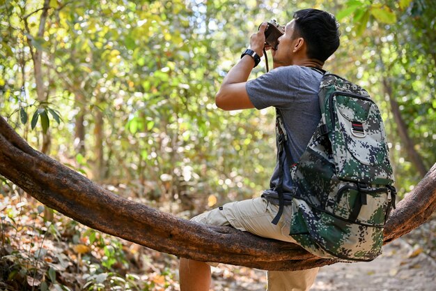 Ein asiatischer Mann mit Trekkingausrüstung, der auf einer Baumrebe sitzt und mit seiner Filmkamera ein Foto macht