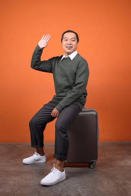 Ein asiatischer Mann winkt mit der Hand und sagt "Hi" zum Abschied, während er auf dem Gepäck sitzt und bereit ist, in den Urlaub zu gehen.