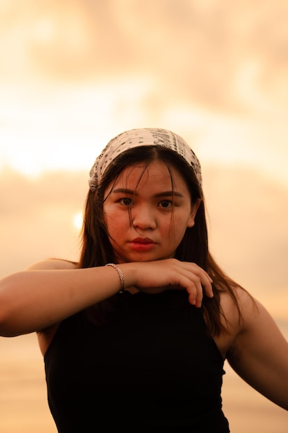 Ein asiatischer Teenager, der ein weißes Bandana und ein schwarzes Hemd mit einem sexy Ausdruck auf dem Strandsand trägt