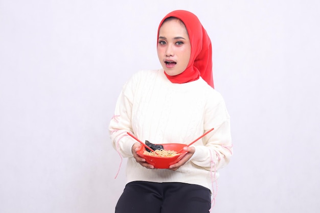 Ein asiatisches Mädchen mit einem Hijab ist schockiert, als es mit beiden Händen eine Schüssel mit Ramen-chinesischem Essen hält