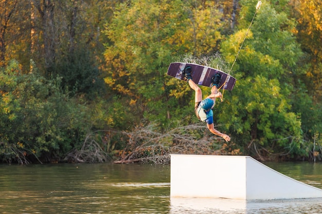 Ein Athlet springt bei Sonnenuntergang von einem Sprungbrett im Wakeboard-Park. Ein Mann führt einen Trick auf einem Brett aus