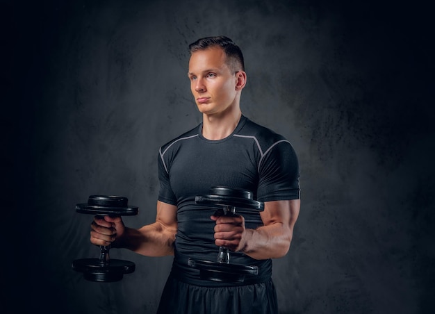 Ein athletischer Fitness-Mann in Sportbekleidung hält eine Reihe von Hanteln auf grauem Hintergrund.