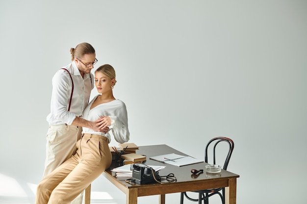 ein attraktives junges Paar in stilvollen Kleidern posiert liebevoll, während sie im Büro in der Nähe des Arbeitstisches eine Affäre haben