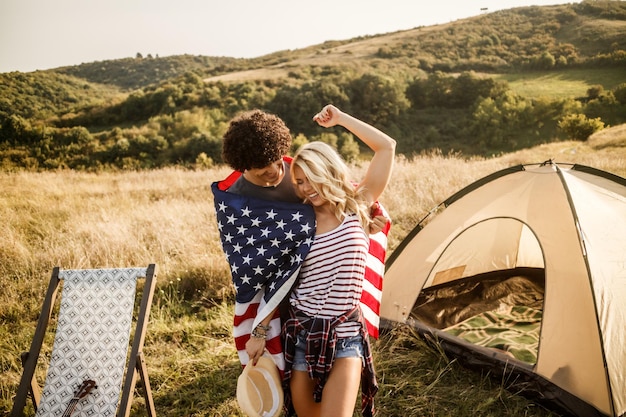 Ein attraktives Liebespaar, das in amerikanische Flagge gehüllt ist und vor einem Zelt auf dem Campingplatz genießt und feiert.