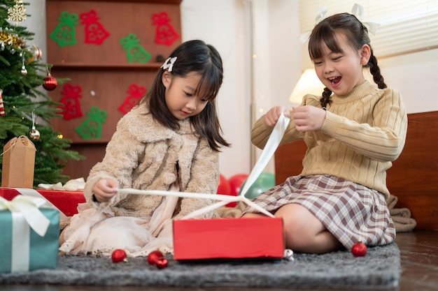 Ein aufgeregtes asiatisches Mädchen öffnet ihr Weihnachtsgeschenk mit ihrer jüngeren Schwester in der Nähe des Weihnachtsbaums