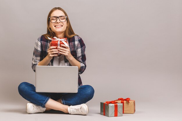 Ein aufgeregtes lässiges Mädchen, das Laptop-Computer hält, während auf einem Boden mit Stapel Geschenkboxen sitzt