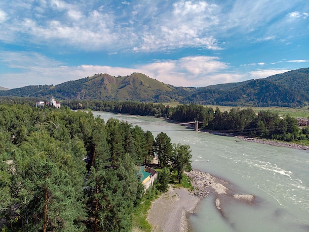 Ein Außenfoto, das von einer Drohne aufgenommen wurde. Blick auf die großen felsigen Berge mit vielen Bäumen und einem fließenden Fluss und mehreren Gebäuden. Waldlandschaft mit Erholungszentrum.