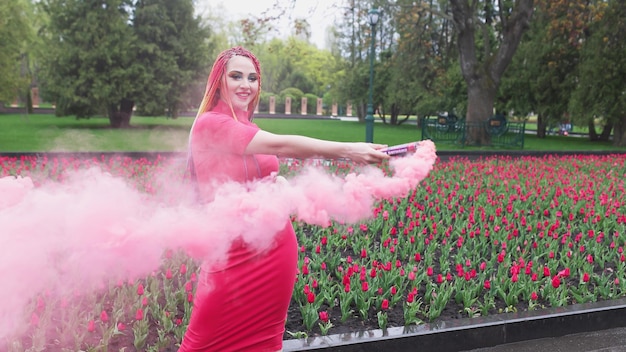Ein außergewöhnliches Mädchen in einem roten Kleid mit Make-up und farbigen Zöpfen. Süß lächeln und posieren in dickem rosa Rauch im Park im Regen