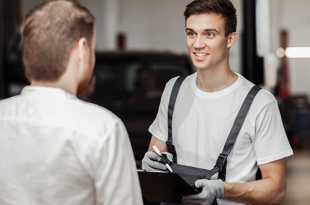 Ein Automechaniker lächelt seinen Kunden an und lädt ihn ein, sich sein Auto anzuschauen.