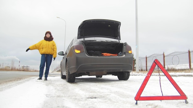 Ein Autounfall, eine junge Frau, die an einem kaputten Auto steht und ein Auto fängt, um Hilfe zu erhalten, ein Notfallschild