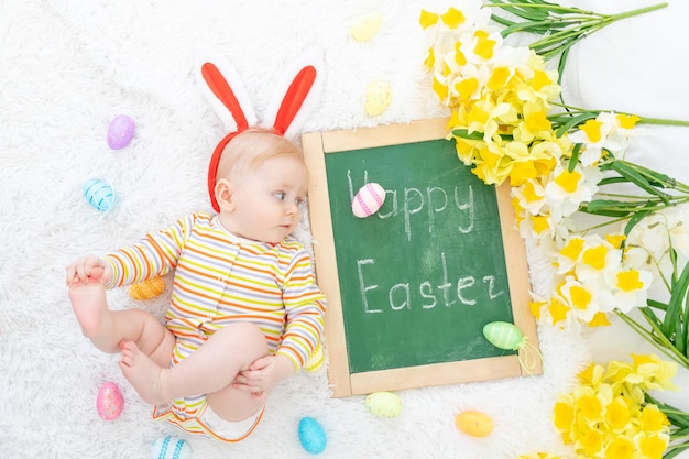 Ein Baby in einem Hasenhut auf einem Bett mit Ostereiern und der Aufschrift fröhliche Ostern