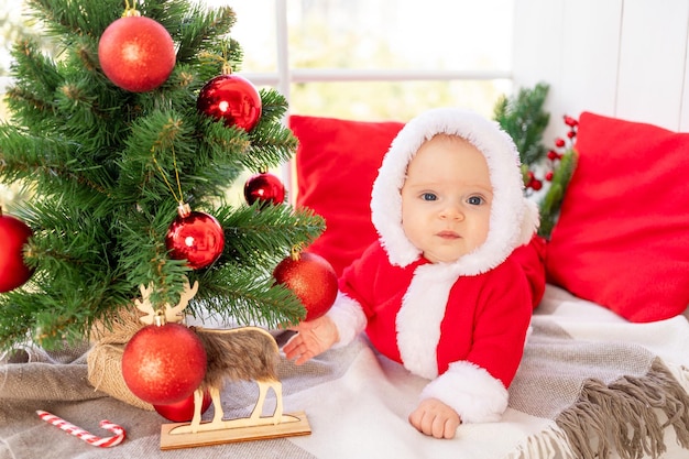 Ein Baby in einem Weihnachtsmann-Kostüm unter dem Weihnachtsbaum spielt mit roten Weihnachtsbällen, die auf dem Fenster des Hauses liegen, das Konzept des neuen Jahres
