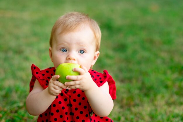 Ein Baby isst einen grünen Apfel in einem roten Bodysuit auf dem grünen Gras im Sommerraum für Text