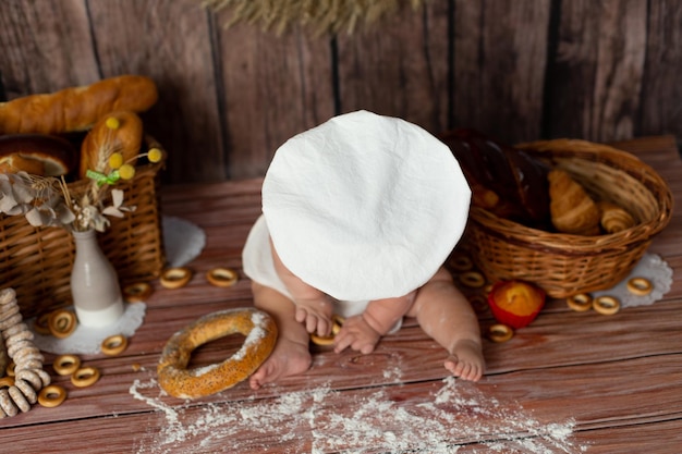 Ein Baby mit weißer Kochmütze sitzt auf einem Tisch mit Brot und Bagel.