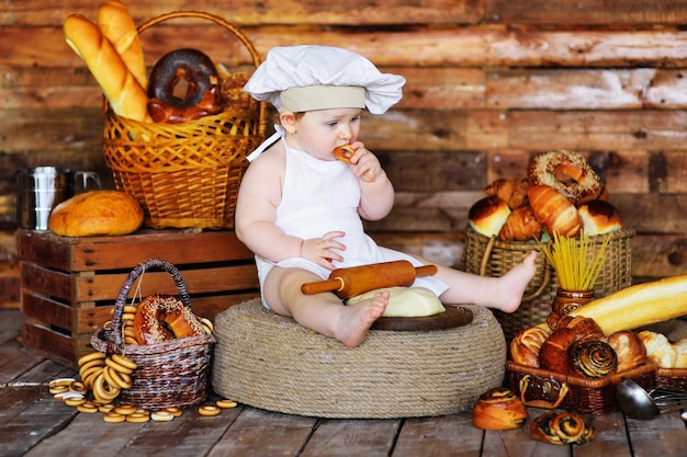 Ein Babybäckerjunge in Kochmütze und Schürze rollt rohen Teig mit einem hölzernen Nudelholz aus und isst einen Bagel vor dem Hintergrund von Backwaren.