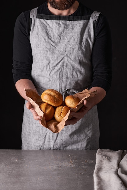 Ein Bäckermann mit Bart in grauer Schürze steht vor schwarzem Hintergrund und hält, bricht, schneidet leckeres, knuspriges Brot, Brötchen, Baguette ab.