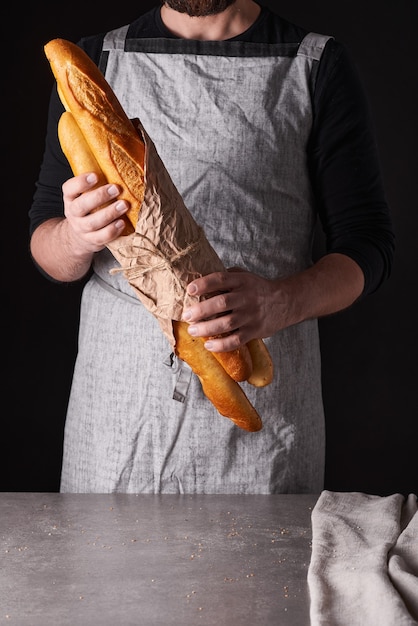 Ein Bäckermann mit Bart in grauer Schürze steht vor schwarzem Hintergrund und hält, bricht, schneidet leckeres, knuspriges Brot, Brötchen, Baguette ab.