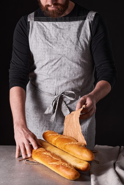 Ein Bäckermann mit Bart in grauer Schürze steht vor schwarzem Hintergrund und hält, bricht, schneidet leckeres, knuspriges Brot, Brötchen, Baguette ab.