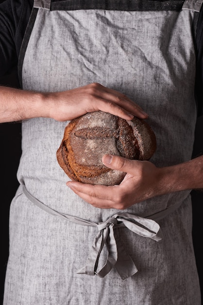Ein Bäckermann mit Bart in grauer Schürze steht vor schwarzem Hintergrund und hält, bricht, schneidet leckeres, knuspriges Brot, Brötchen, Baguette ab.