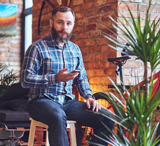 Ein bärtiger Hipster-Mann in einem Fleece-Shirt mit Smartphone in einem Raum mit Loft-Interieur.