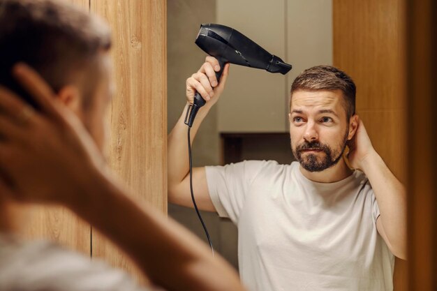 Ein bärtiger Mann betrachtet sich in einem Badezimmer im Spiegelbild und trocknet sein Haar