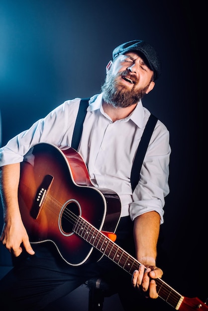 Ein bärtiger Mann in Anzug und Hut sitzt und spielt auf der Akustikgitarre. Studioporträt