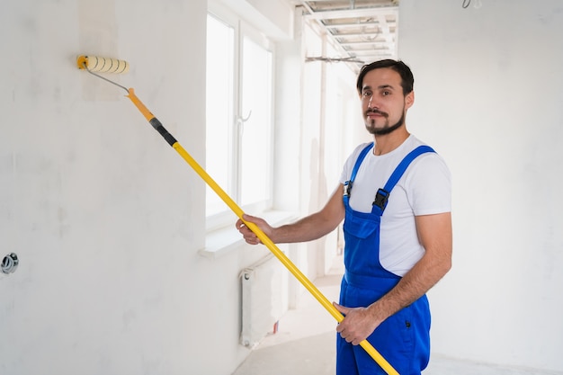 Ein bärtiger Mann in einem blauen Overall malt eine Wand im Flur. Er benutzt eine Walze