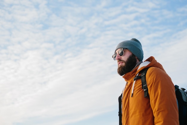 Ein bärtiger Mann mit Rucksack in Sonnenbrille und warmem Hut vor dem Hintergrund des blauen Himmels Das Konzept des Wanderns und des aktiven Lebensstils
