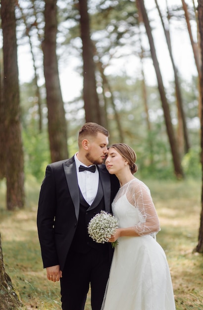 Ein bärtiger, stilvoller Bräutigam im Anzug und eine schöne blonde Braut in einem weißen Kleid mit einem Strauß in den Händen stehen und umarmen sich in der Natur im Kiefernwald.