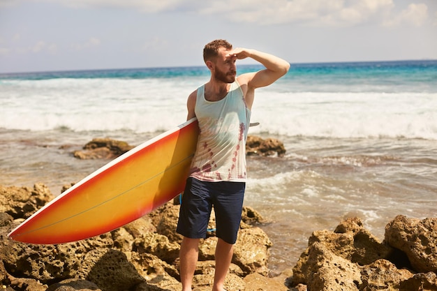 Ein bärtiger Surfer mit einem Surfbrett geht am Strand spazieren und beobachtet die Wellen vor Ort