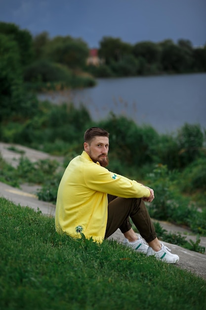 Ein bärtiger Typ sitzt bei Sonnenuntergang in einer gelben Jacke in der Nähe des Sees auf dem Gras