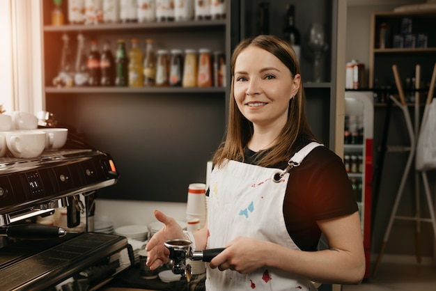 Ein Barista mit weißer Schürze lächelt und posiert mit einem Metallstampfer und einem Siebträger mit Kaffee