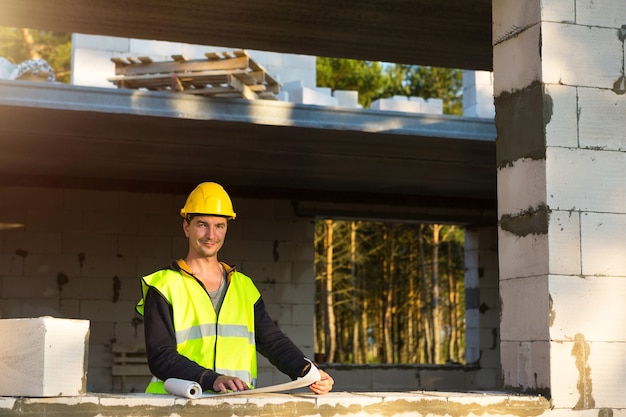 Ein Bauingenieur und Designer in einem gelben Bauarbeiterhelm studiert eine Zeichnung eines Gebäudes auf einer Baustelle. Ein Haus aus Porenbetonsteinen, ein Arbeiter dabei an Fenster und Wand