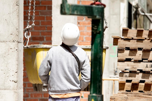 Ein Baumeister in einem harten Hut beobachtet das Anheben eines Behälters mit einer Betonmischung an einer Kette