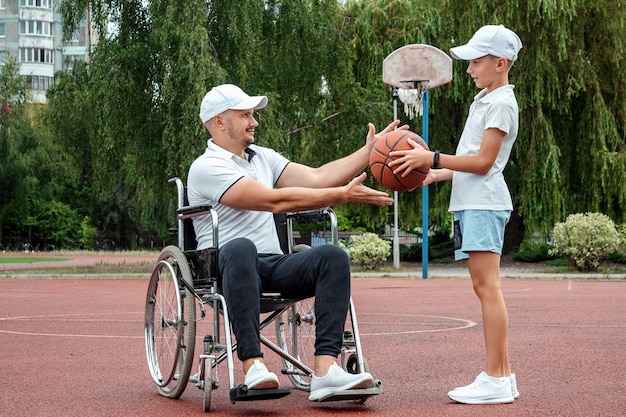 Ein behinderter Vater spielt mit seinem Sohn auf der Straße. Rollstuhlkonzept, behinderter Mensch, erfülltes Leben, Vater und Sohn, Aktivität, Fröhlichkeit.