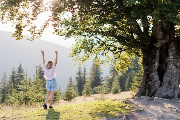 Ein bezaubernder malerischer Blick auf die Berge Eine junge Frau steht in der Nähe eines großen Baums auf einem Hügel und hob die Hände in die Karpaten