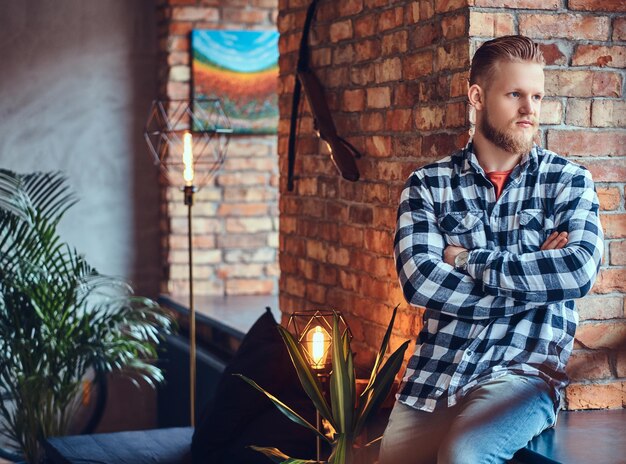 Ein blonder bärtiger Hipster-Mann in Jeans und Fleece-Shirt, der in einem Raum mit Loft-Interieur posiert.