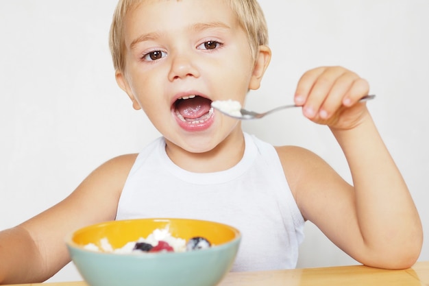 Ein blonder Junge mit braunen Augen in einem weißen T-Shirt isst Hüttenkäse mit Beeren an einem Holztisch.