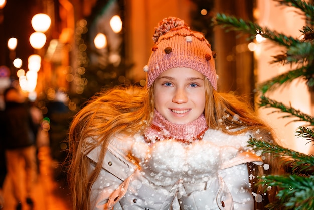 Ein blonder Teenager mit langen Haaren in Hut und Winterkleidung an einem festlichen Winterabend auf der Str...