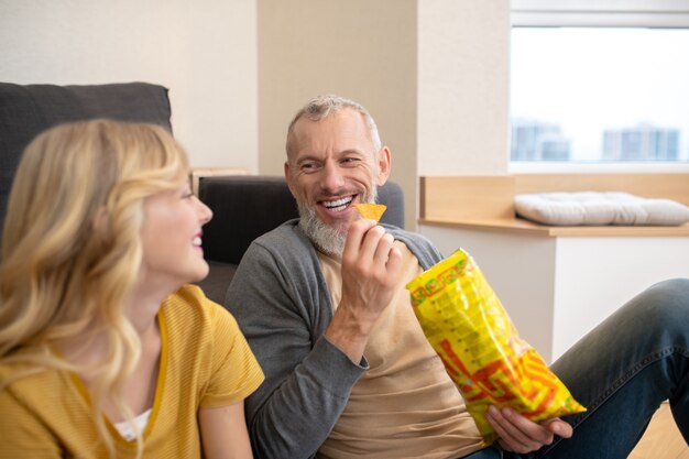 Ein blonder Teenie sitzt mit ihrem Vater auf dem Boden und isst Chips