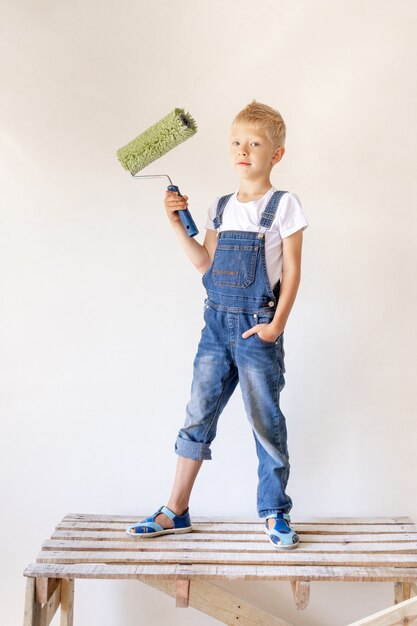 Ein blondes Kind steht auf einer Bauleiter in einer Wohnung mit weißen Wänden und einer Walze in den Händen, einem Ort für Text, dem Konzept der Reparatur