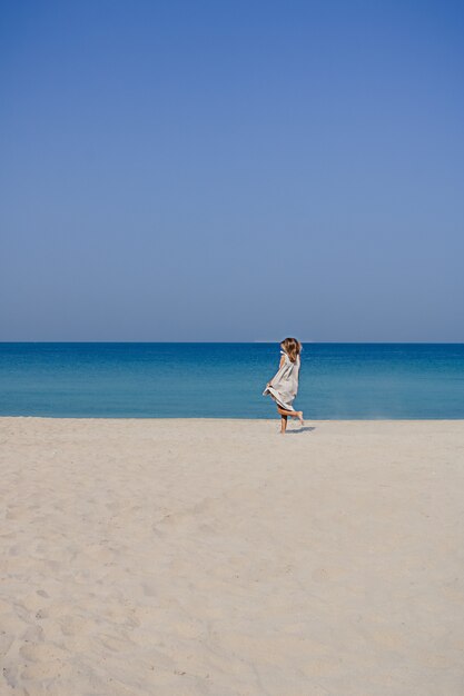 Ein blondes lächelndes Mädchen in einem Maxi-Leinenkleid mit flatternden Haaren, die auf einem Sandstrand springen und tanzen