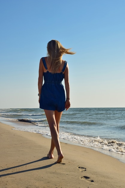 Ein blondes Mädchen in Denim-Overalls geht am Sandstrand entlang. Rückansicht